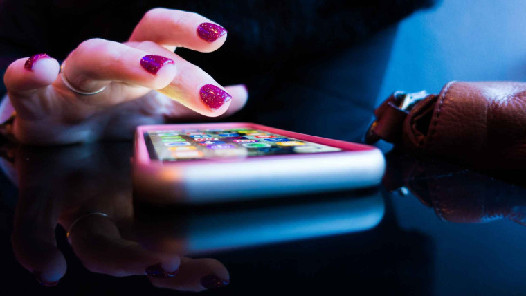 A close-up photo of hands using a smartphone, viewed at an angle on a reflective surface.